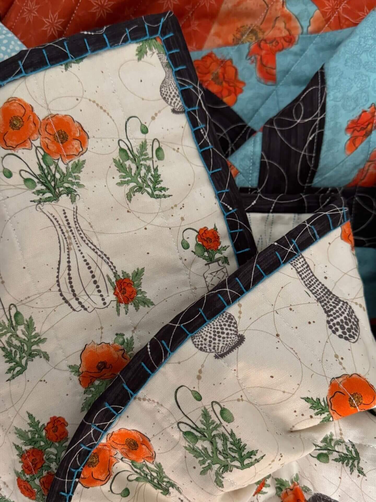 Close-up of a folded quilt corner, showing the dark binding fabric stitched with bright blue thread in the big stitch technique. The quilt fabric has a white background with orange poppies and a vase outline.